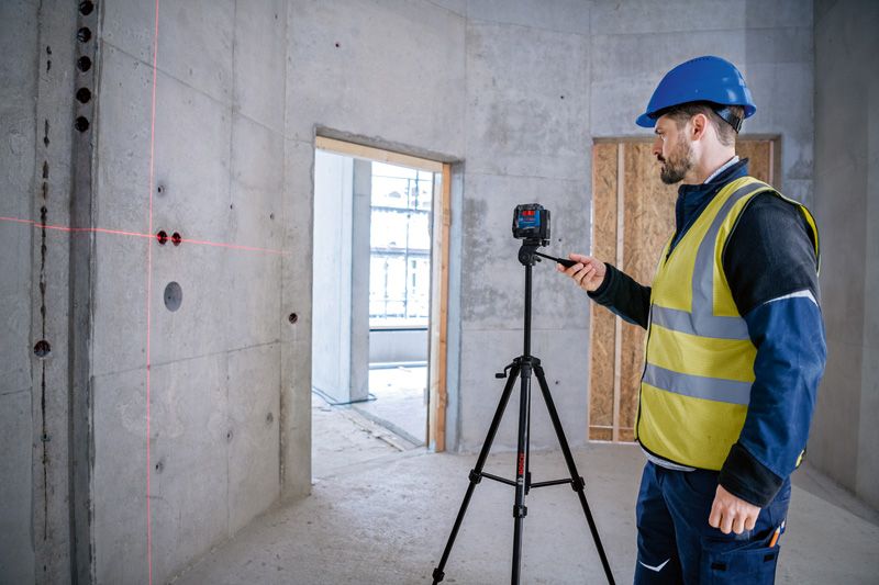 A person wearing safety equipment adjusts a laser leveling tool on a tripod in a concrete room.