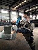 A person wearing safety equipment uses a straight grinder on a metal sheet.