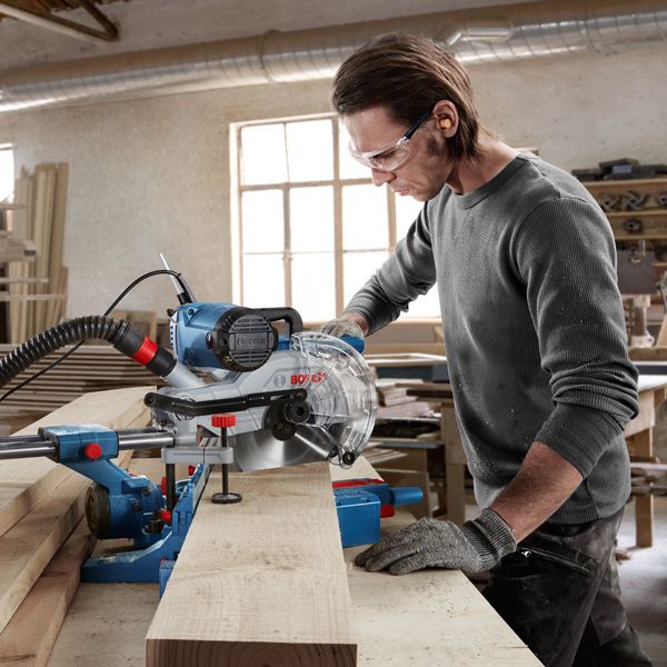A person wearing safety equipment uses a miter saw to cut a wooden board in a workshop.