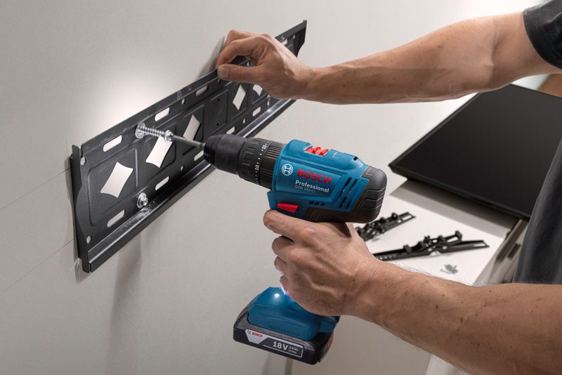 A person drills screws into a wall mount bracket using a cordless power drill.