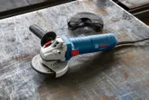 Angle grinder with side handle and detached wheel guard on a scratched metal work surface.