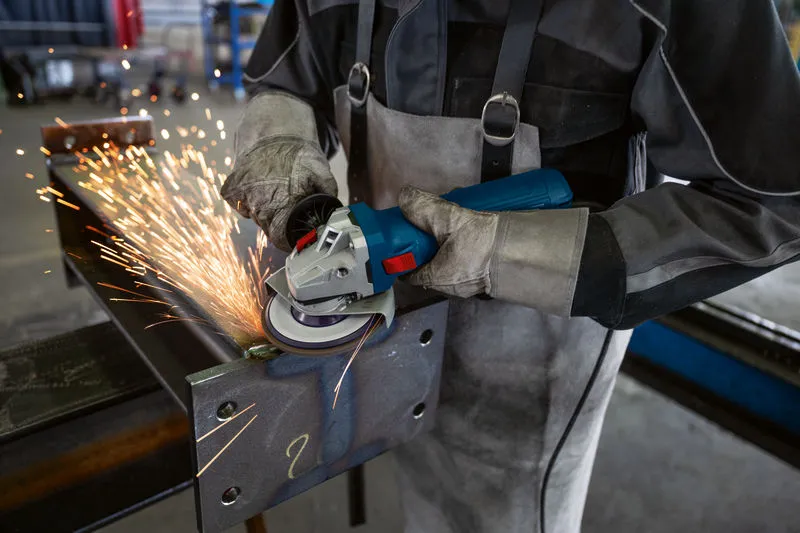 A person wearing safety equipment grinds a steel beam with a power tool, producing sparks.