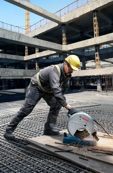 A person wearing safety equipment cuts metal pipes at a construction site with a cut-off saw.