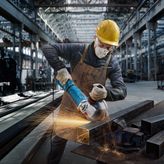 A person wearing safety equipment grinds a metal beam in an industrial workshop.