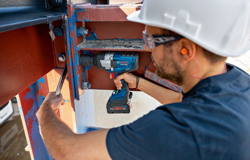 A person wearing safety equipment uses a cordless impact wrench on a steel beam.