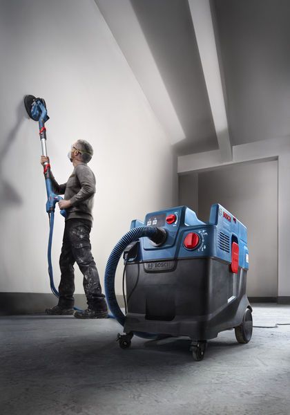 A person wearing safety equipment sands a wall using a drywall sander, connected to an extractor.