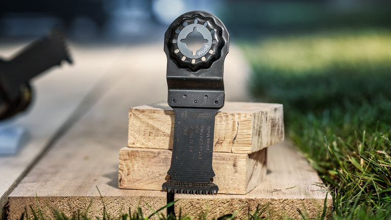 Oscillating multi-tool blade resting on stacked wooden planks outdoors.