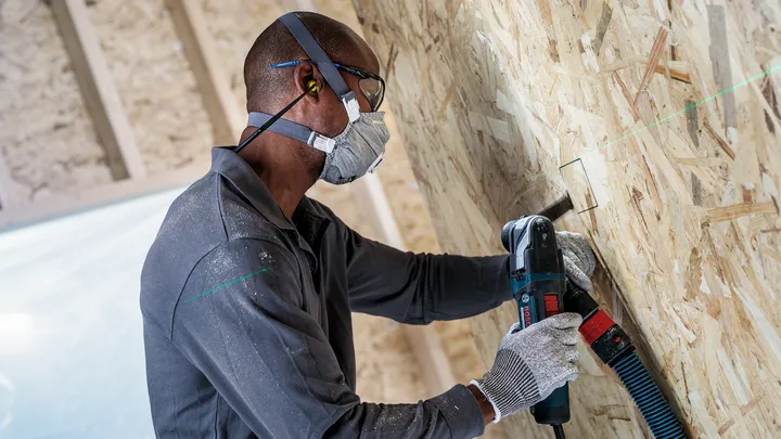 Person wearing safety equipment drills a rectangular hole in a wooden wall.