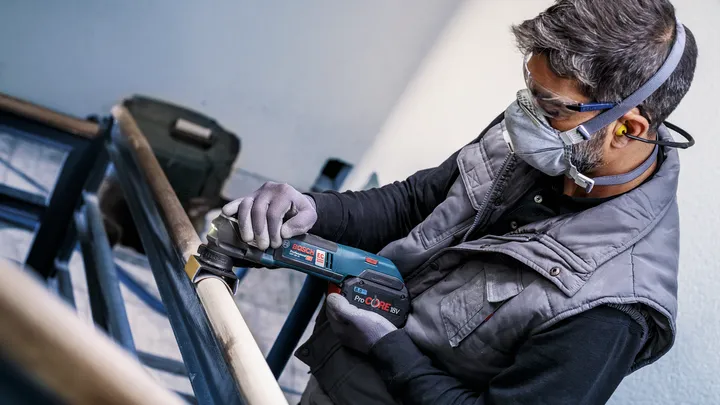 Person wearing safety equipment sands a wooden railing with a cordless power tool.