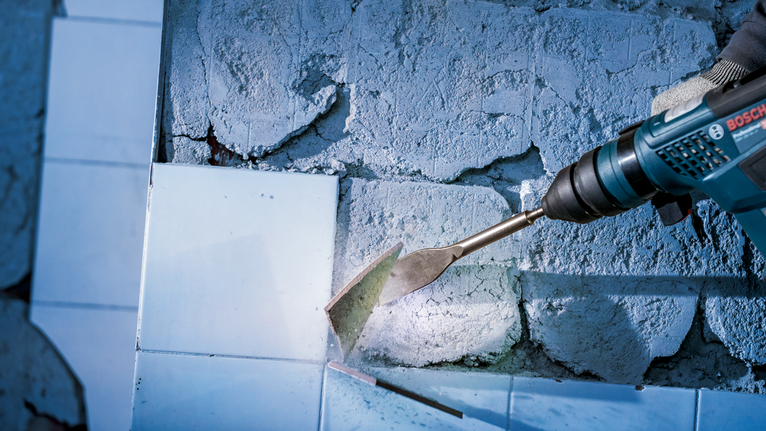 Person wearing safety equipment uses a power tool to remove tiles from a wall.