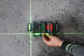 A person adjusts a laser leveling tool projecting green lines on a concrete floor.