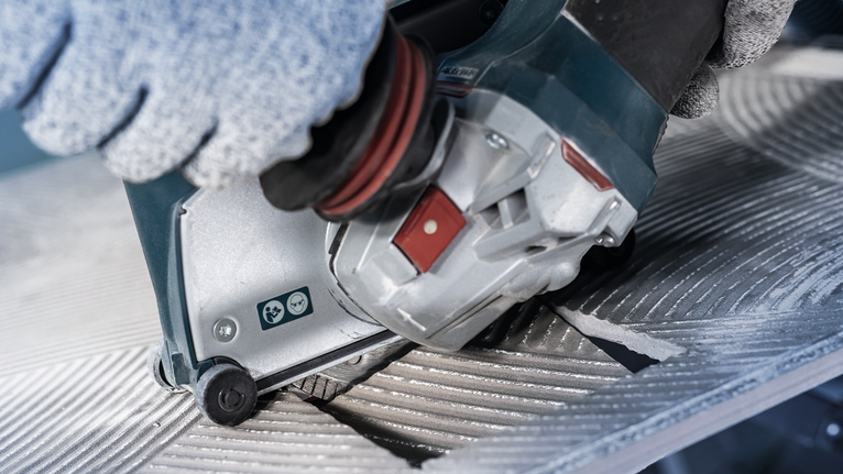 Person wearing safety equipment operates a power tool to cut a grooved metal surface.