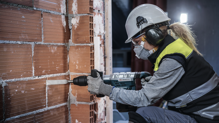 A person wearing safety equipment drills into a brick wall with a power tool.