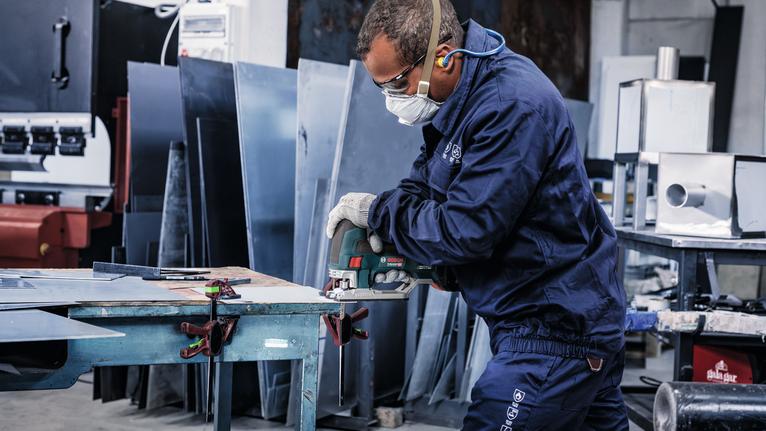A person wearing safety equipment cuts sheet metal with a power tool in a workshop.