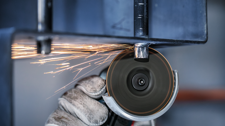 Person wearing safety equipment uses an angle grinder to cut metal, producing sparks.
