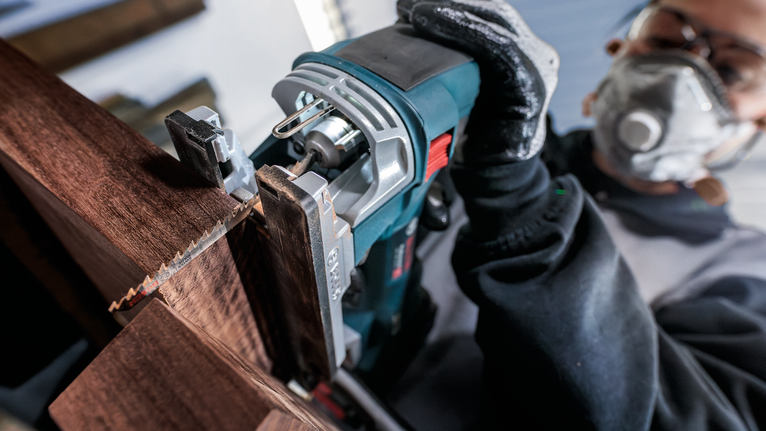 Person wearing safety equipment cuts wood with a handheld jigsaw.