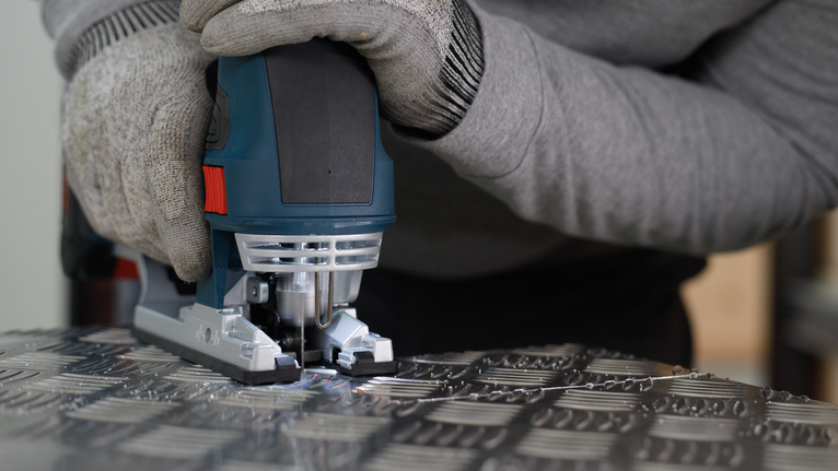 Person wearing safety equipment guides a jigsaw to cut patterned metal sheet.