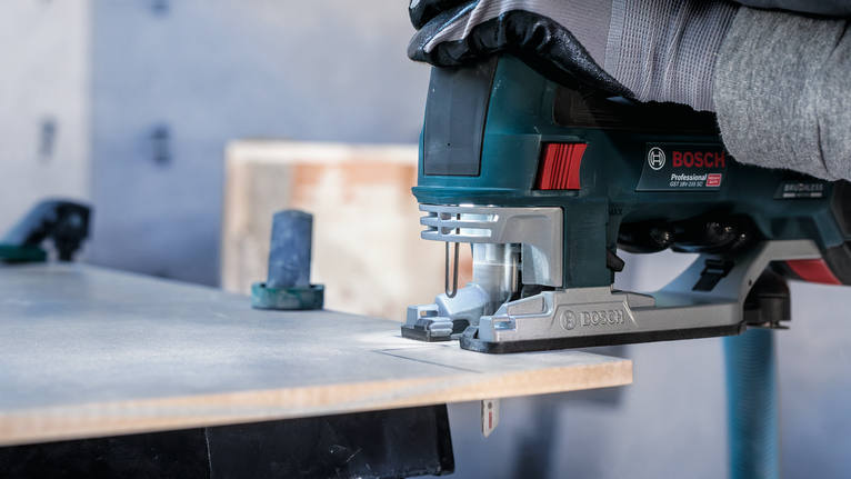Person wearing safety equipment cuts wood with a power jigsaw.