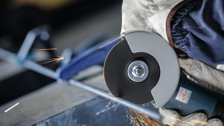 Person wearing safety equipment grinds a metal bar with a cutting tool.