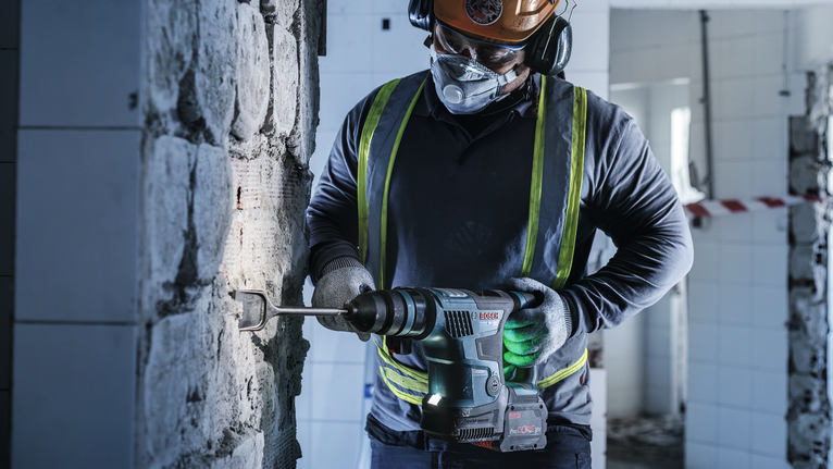 A person wearing safety equipment uses a rotary hammer to chisel a wall.