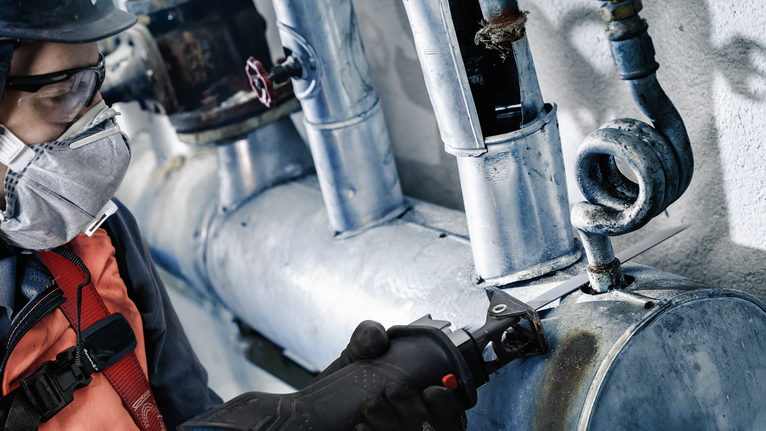 A person wearing safety equipment cuts a metal pipe with a reciprocating saw.