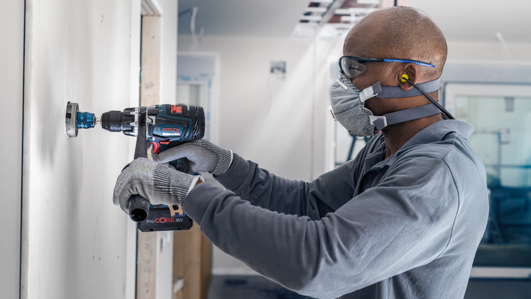 Person wearing safety equipment uses a power drill to cut into a wall.