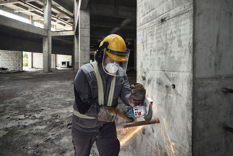 A person wearing safety equipment grinds rebar on a concrete wall with a cordless angle grinder.