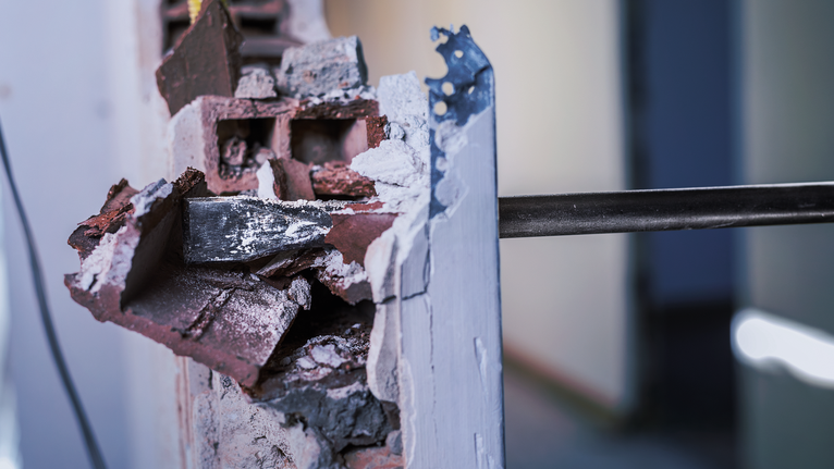 A rotary hammer demolishes a concrete wall, exposing broken bricks.