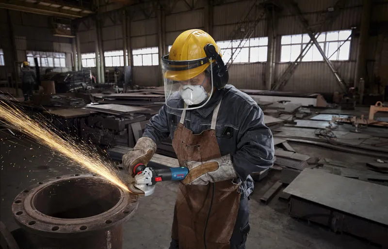 A person wearing safety equipment uses an angle grinder to cut metal, producing sparks.