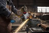 A person wearing safety equipment grinds a metal pipe, producing bright sparks.
