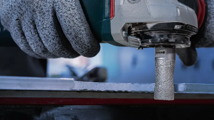 Person wearing safety equipment uses a rotary tool with a textured bit on a metal surface.
