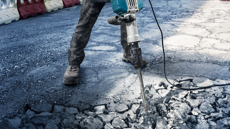 A person wearing safety equipment breaks concrete with a heavy-duty demolition tool.
