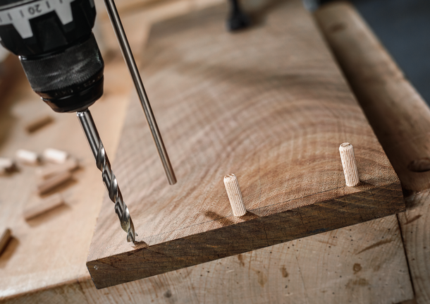 Drill making a hole in a wooden board with dowels inserted nearby.