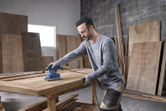 A person wearing safety equipment sands a wooden tabletop in a workshop.