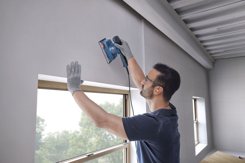 A person wearing safety equipment sands a wall near a window using an orbital sander.