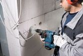A person wearing safety equipment drills a circular hole into a concrete wall.