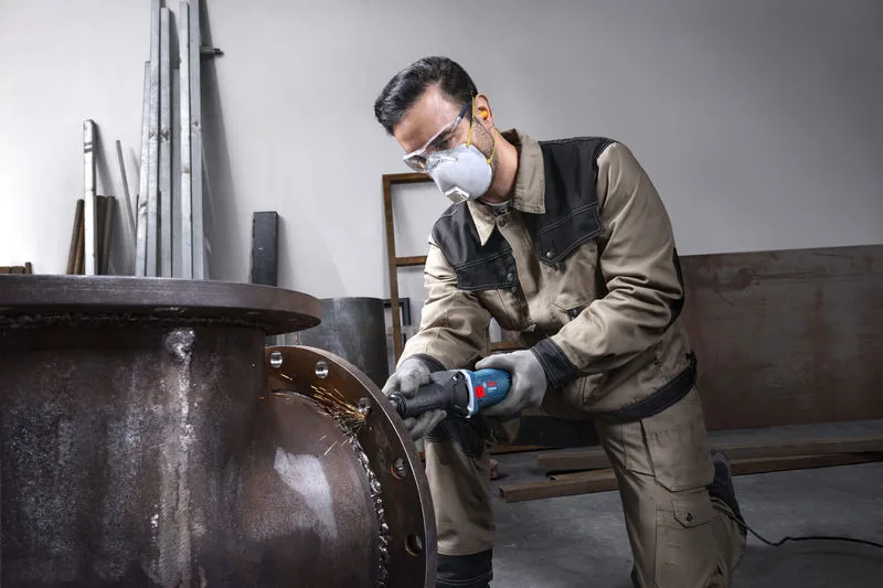 A person wearing safety equipment grinds the edge of a large metal pipe.