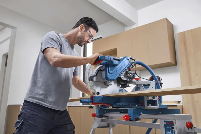 A person wearing safety equipment cuts wood with a mitre saw in a workshop.