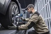 A person wearing safety equipment uses a cordless impact wrench to tighten a car wheel.