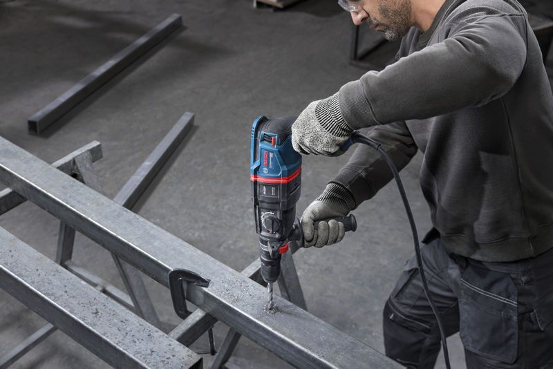 A person wearing safety equipment drills into a metal beam with a rotary hammer.