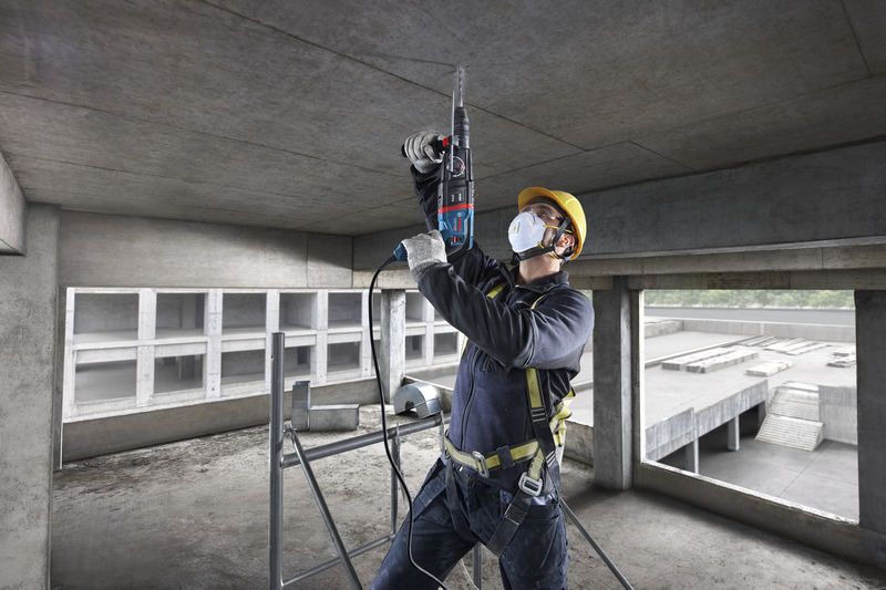 A person wearing safety equipment drills into a concrete ceiling with a rotary hammer.