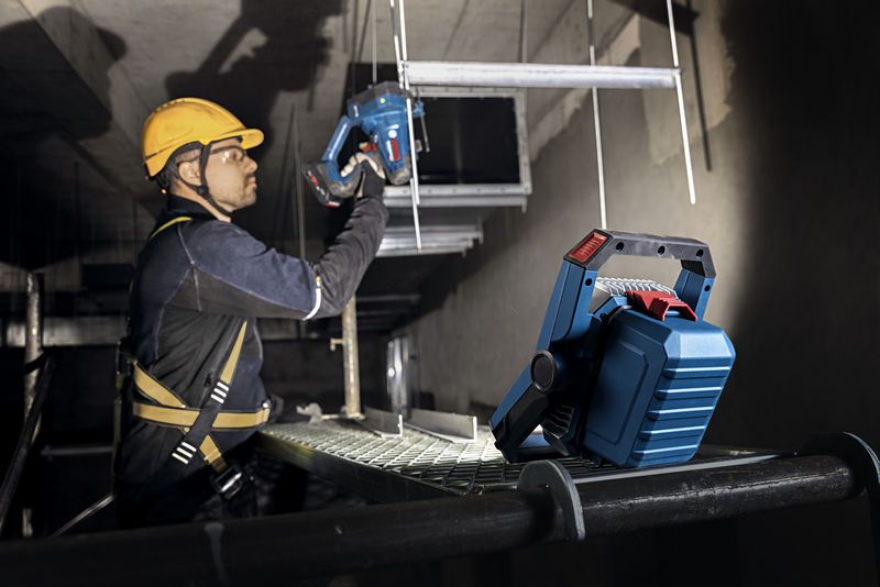 A person wearing safety equipment uses a cordless power tool under a bright jobsite light.