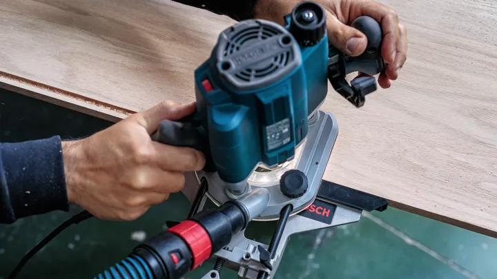 Person guides a power tool with dust extraction while shaping the edge of wood.