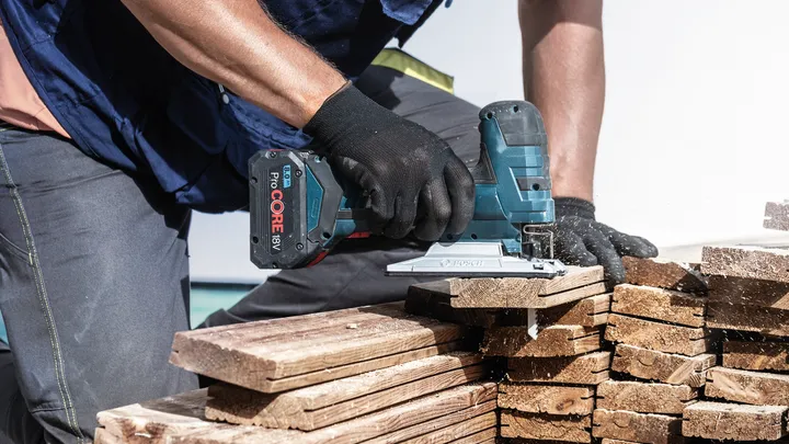 Person wearing safety equipment uses a cordless jigsaw to cut stacked wooden planks.