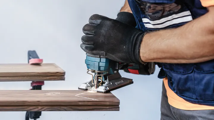Person wearing safety equipment cuts wood with a jigsaw.