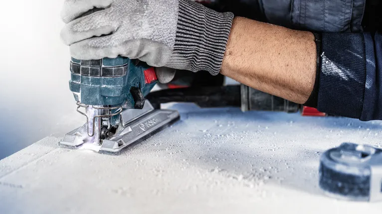 Person wearing safety equipment uses a jigsaw to cut a sheet of material.