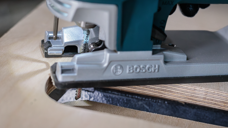 A person wearing safety equipment guides a jigsaw to cut a curved shape in plywood.