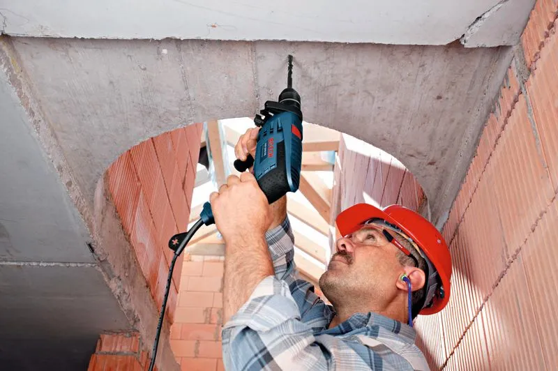 A person wearing safety equipment drills into a concrete ceiling using an impact drill.