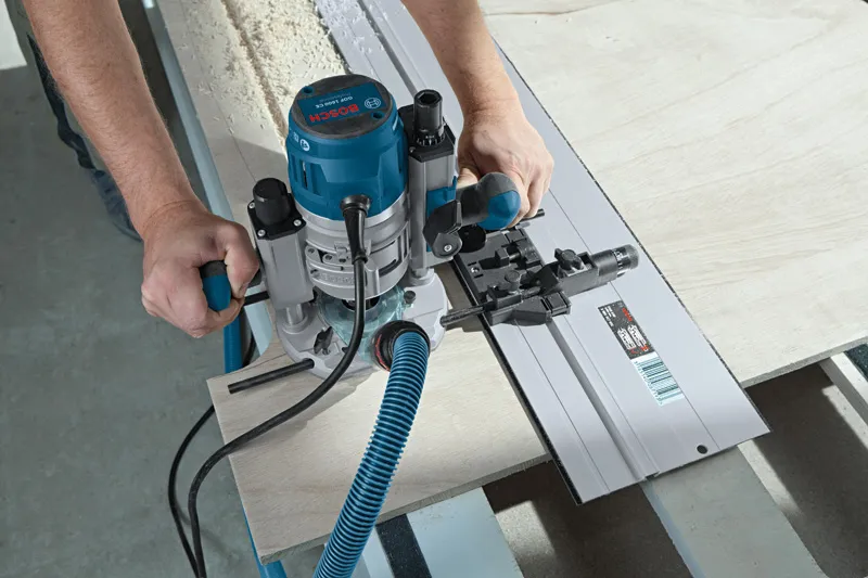 A person uses a blue router to cut wood along a straight guide rail.
