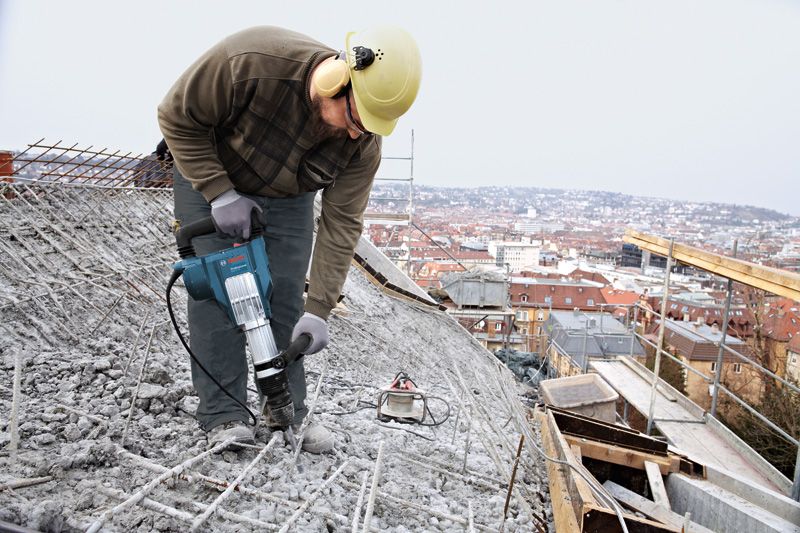 Asmuo, dėvintis saugos įrangą, griovimo plaktuku skaldo betoną ant stogo.